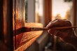 © sogap - A persons hand holding a paintbrush applies stain to a wooden window frame.