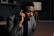 © Viktor - Young black man wears headset talks into microphone uses laptop computer at home desk in evening. Focused pro works remotely during night indoors. Serious person discusses project via online call.