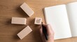 © Christine - Focused hand writing down strategic goals and plans in a blank notebook next to wooden blocks on a wooden desk.