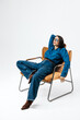 © wedmoments.stock - A model wearing a shirt and jeans sits on a modern chair in a minimalist studio setting, showcasing contemporary fashion and a relaxed pose.