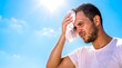 © Obbinokes - Young man wiping sweat with a towel under intense scorching sun. Portrait of person suffering from heatstroke and high temperature during summer day. Blue sky background with bright sunlight.