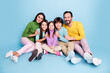 © deagreez - Happy family sitting together on blue studio background, parents and two children smiling and embracing in colorful casual clothes