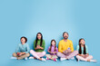 © deagreez - Happy modern family of five sitting cross‑legged on a blue background, parents and children smiling and using smartphones together