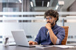 © Tetiana - A thoughtful young Indian man is sitting at a desk in the office and looking at the laptop screen