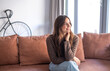 © luciano - A smiling young woman sits on a plush couch in a cozy, modern living room, looking out the window as soft natural light brightens the space and creates a relaxed, content atmosphere.