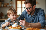 Father, son share waffles with whipped cream at kitchen table. Young boy eats dessert with fork, dad watches with smile. Happy family bonding moment over sweet homemade breakfast, enjoying food