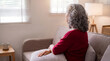 © Mary - Thoughtful elderly woman with gray hair sits alone on a cozy sofa, gazing out a bright window at home.