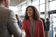 © EricMiguel - Smiling businesswoman with curly dark hair in coral blazer warmly shaking hands in bright modern office lobby, wearing lanyard and grey sweater