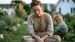 © forenna - Mother in muddy gardening clothes sitting cross-legged on backyard lawn in full parental collapse, toddler happily digging nearby, wide shot of overgrown summer garden under