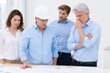 © Дмитрий Баронин - Group of professionals analyzing architectural plans on a table in a bright office space, featuring a man in a hard hat, two men in blue shirts, and a woman in a white blouse