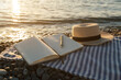 © Evgeniia Primavera - Open notebook with pen resting on striped beach blanket near calm sea water at golden sunset, straw hat beside journal creating peaceful summer vacation atmosphere, travel lifestyle concept