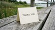 © Mudassir - White folded paper card with the text thank you printed on it sitting on a weathered wooden boardwalk railing outdoors