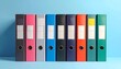 © raya - A row of colorful, upright binders. Each is a different shade, with a blank label. They stand against a pastel blue backdrop