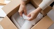 © Viktor - Person carefully packs item in bubble wrap into cardboard box. Preparing fragile goods for shipping. Secure product for delivery. Hands protect contents inside package.