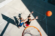 © Studio Marmellata - Two young athletes compete in a basketball game on an outdoor court. One jumps to shoot the ball, while the other defends, attempting to block the shot near the hoop.