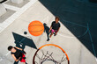 © Studio Marmellata - Two diverse women play basketball on an outdoor court. A basketball is in mid-air, heading towards the hoop. Both women look up, enjoying their game under the sun.