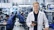 © ArtFocus Studio - Smiling Young Male Engineer in White Lab Coat Holding Tablet in Modern Electronics Manufacturing Facility with Coworkers at Workstations in the Background