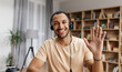 © Prostock-studio - Cheerful arab man in headset having web conference, waving hand at camera, gesturing hello and smiling, enjoying online communication, webcam view
