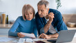 © Prostock-studio - A couple sits together at a table looking at bills and important documents. They are focused and discussing details. A laptop and receipts are present on the table.
