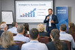 © vefimov - Businessman speaks to audience during workshop presentation. Presenter gestures toward a growth chart on projection screen. Attendees sit facing speaker, focused on session content