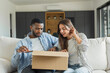 © Daniel - Happy multiracial couple sitting on a sofa, opening and looking inside a delivered cardboard box, enjoying online shopping delivery in their modern living room