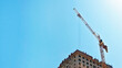 © Irina - Construction crane lifting materials at a high-rise building under construction against a clear blue sky, showcasing modern architecture and urban development, copyspace