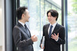 © kapinon - 新緑のオフィスの窓際で会話する若手ビジネスマンと上司、取引先のコミュニケーション商談と成長の仕事イメージ