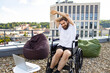 © sofiko14 - A smiling man in a wheelchair stretches his arms on a rooftop patio with beanbag chairs and a laptop nearby, overlooking a city skyline