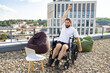 © sofiko14 - A man in a wheelchair raises his arm in a gesture of triumph while sitting on a rooftop terrace with a laptop and beanbag chairs
