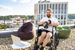 © sofiko14 - A smiling man in a wheelchair is lifting dumbbells on a rooftop terrace with a city view, accompanied by beanbag chairs and a laptop