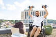 © sofiko14 - A smiling man with a beard sits in a wheelchair on a rooftop terrace, lifting dumbbells overhead while looking at the camera