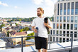 © sofiko14 - A man with a beard and tied-back hair holds a water bottle and a rolled-up yoga mat while standing on a rooftop overlooking a city