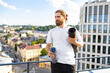 © sofiko14 - A bearded man with his hair tied back holds a yoga mat and water bottle while standing on a rooftop overlooking a city