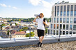 © sofiko14 - A man with long hair tied back drinks from a water bottle while holding a yoga mat on a rooftop overlooking a city