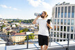© sofiko14 - A man with long hair and a beard drinks from a water bottle while holding a yoga mat on a rooftop overlooking a city