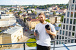 © sofiko14 - A bearded man in a white t-shirt holds a yoga mat and water bottle while standing on a rooftop overlooking a city