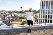 © sofiko14 - A man with a beard and headphones around his neck holds a water bottle and a selfie stick while taking a photo on a rooftop overlooking a city