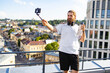 © sofiko14 - A bearded man in athletic wear holds a water bottle and a selfie stick while recording a video on a rooftop overlooking a city