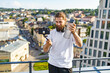 © sofiko14 - A man with a beard and headphones films himself with a smartphone on a selfie stick while holding a water bottle on a rooftop overlooking a city