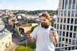 © sofiko14 - A bearded man with headphones around his neck records himself on a smartphone attached to a selfie stick while holding a large water bottle on a rooftop overlooking a city