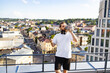 © sofiko14 - A man with headphones around his neck drinks from a metal water bottle while holding a selfie stick with a smartphone attached, overlooking a city skyline
