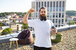 © sofiko14 - A bearded man with headphones around his neck flexes his bicep while holding a water bottle on a rooftop overlooking a city