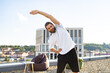 © sofiko14 - A man with a beard and ponytail smiles while stretching his body on a rooftop with a city view and modern building in the background