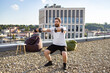 © sofiko14 - A man with a beard and tied-back hair performs dumbbell squats on a rooftop terrace with a city view