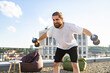 © sofiko14 - A man with a beard and bun hairstyle performs dumbbell exercises on a rooftop with a city view and beanbag chairs