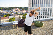 © sofiko14 - A man with a beard and bun hairstyle performs a shoulder stretch on a gravel rooftop with a city view and modern buildings in the background