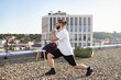 © sofiko14 - A man with a beard and bun hairstyle performs a lunge exercise on a gravel rooftop with a city skyline in the background