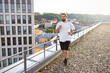 © sofiko14 - A fit man with a beard and long hair runs on a gravel rooftop terrace with a modern building and cityscape in the background during golden hour