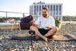 © sofiko14 - A man with a beard sits cross-legged on a yoga mat on a rooftop, eating a salad from a container while filming himself on a smartphone