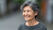 © Nataliia_Trushchenko - Elderly Asian woman with gray hair smiles warmly while standing outdoors in a blurred urban setting, wearing a cozy gray sweater and showcasing natural beauty